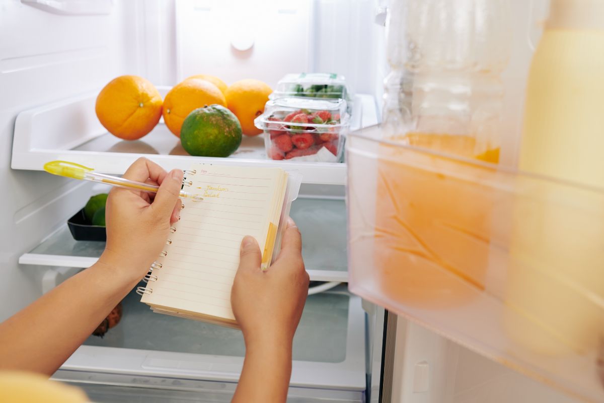 The photo shows a girl's hand and writing on the paper with the fruits inside a fridge which means doing a weekly eat-first inventory helps avoid throwing out food that gets forgotten in the back.