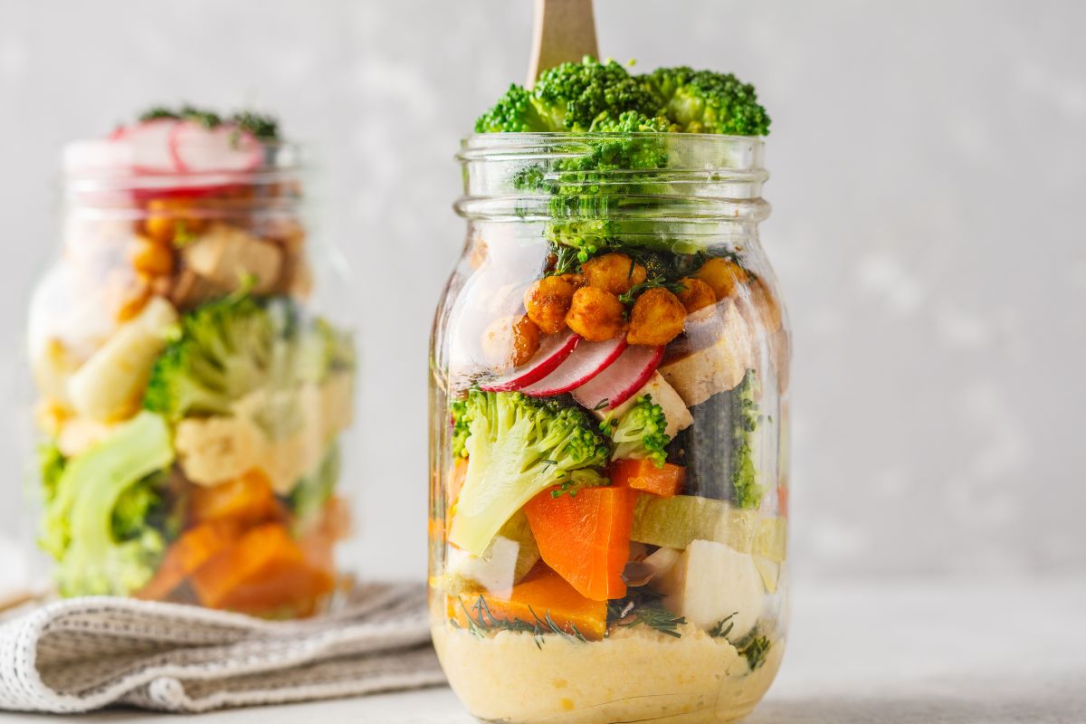The photo shows jars with cut vegetables inside referring to the use of mason jars for salad and leftovers can help save money and prevent waste.