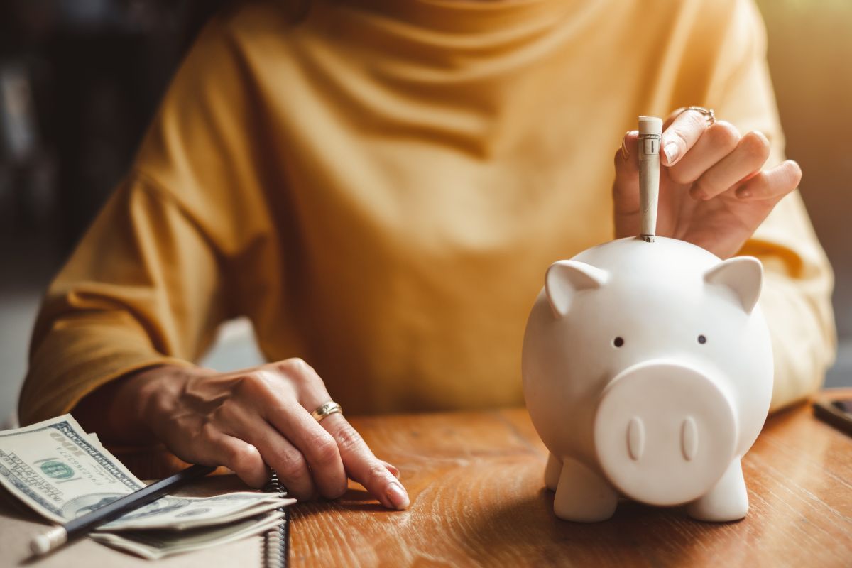 The picture shows a notebook, dollar bills, a pen, and a girl putting a dollar bill into the piggy bank which means breaking it down into smaller chunks keeps you motivated.
