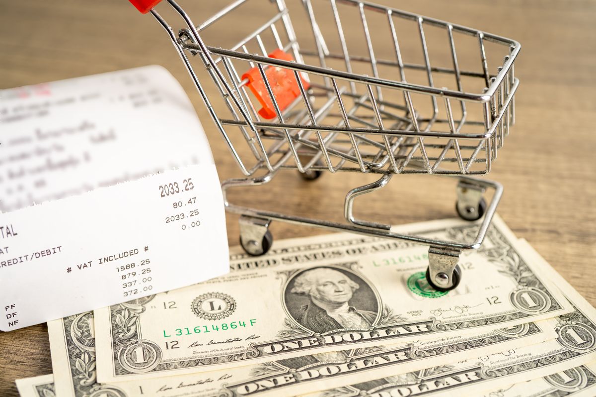 The image shows a cart, dollar bills, and a receipt on the table which means that cutting grocery costs can save you $50 or more each week.