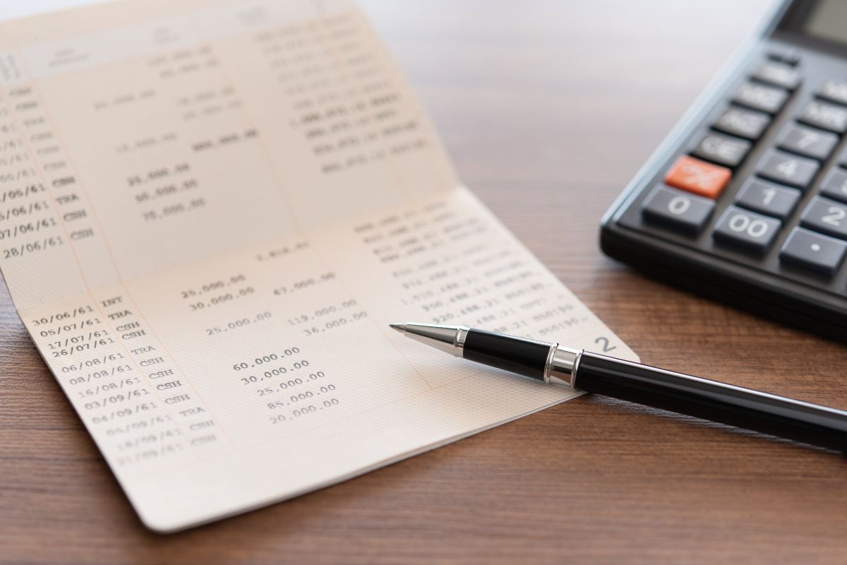 The photo shows a calculator, a pen, and a bank book referring to checking your bank accounts lets you see recent transactions and ensures everything looks alright.