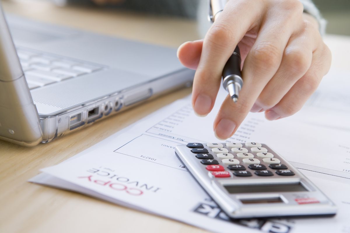 The photo shows a laptop, papers with notes, and a girl's hand using a calculator to review upcoming bills keeps your budget organized and prevents last-minute stress.