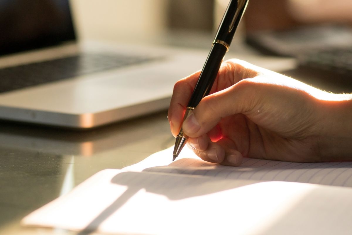 The photo tells about a laptop and a man's hand and writing on the notebook indicating to set a daily financial intention helps you take small but meaningful steps toward financial security.