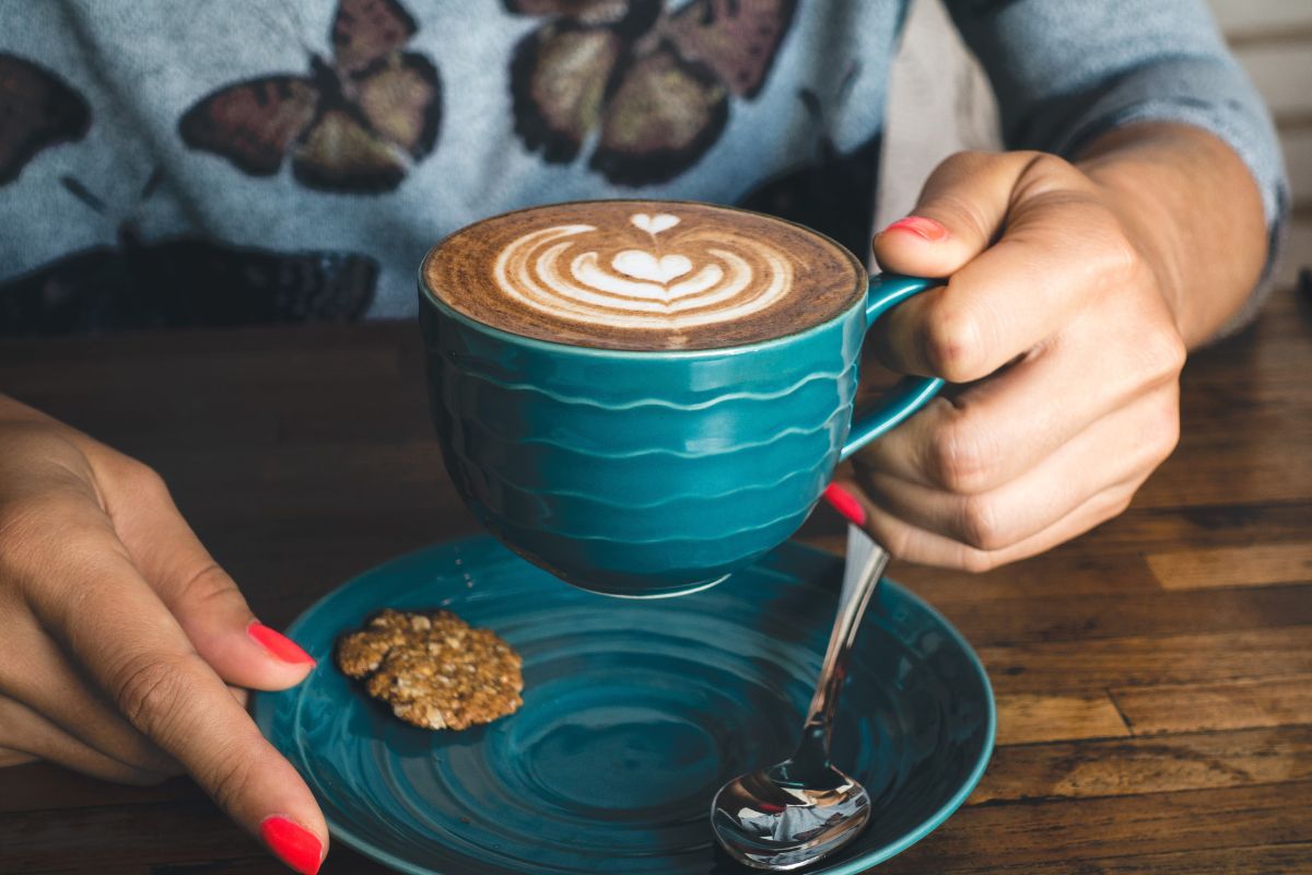 The image tells about a girl's hand holding a plate with a spoon and sugar while sipping an expensive coffee which means optimizing your morning coffee habit is an easy way to cut back on unnecessary spending without feeling deprived.