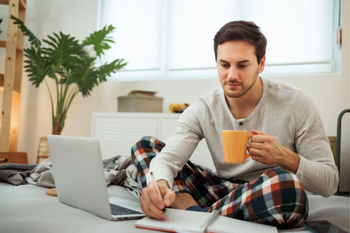 The image tells about a laptop and a man writing on the paper while having a coffee referring to checking out the billionaire's morning routine helps you stay on top of your money and work toward financial freedom.