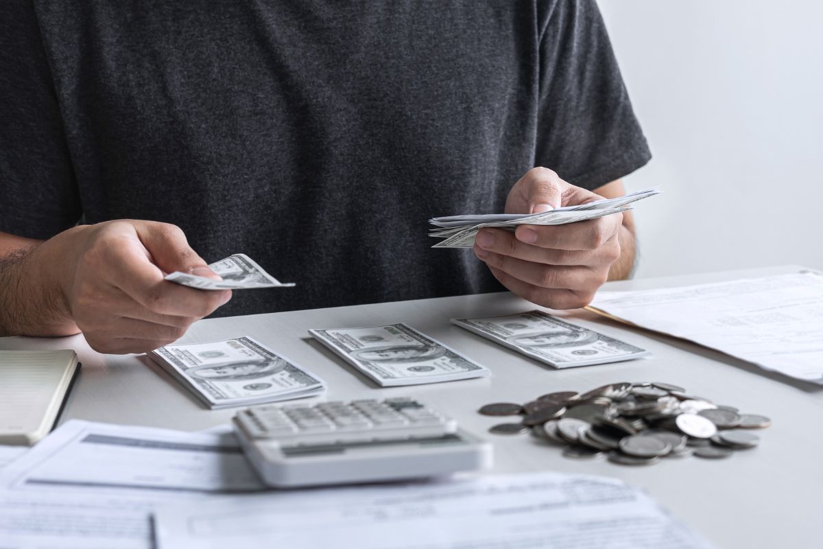The picture shows papers, coins, a calculator, and a man grouping the dollar bills which refers to tracking every dollar to help find wasteful spending and cut unnecessary costs.