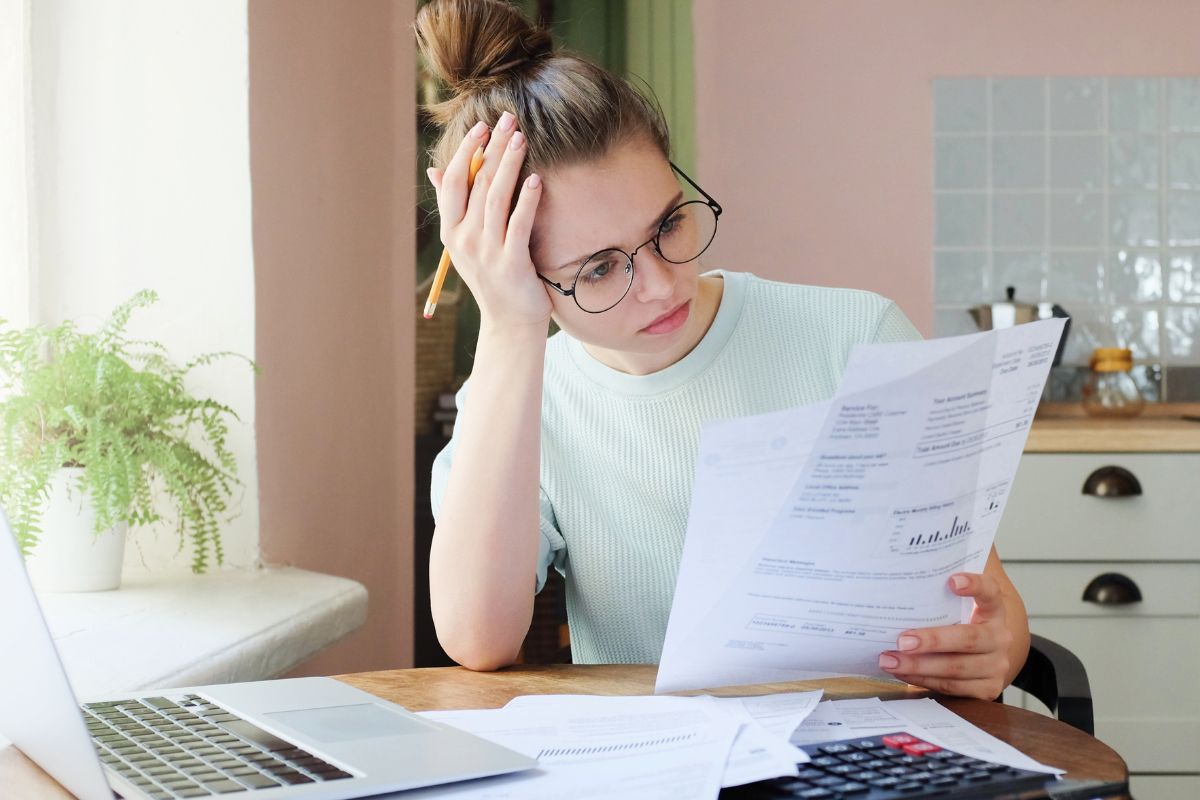 The image of a laptop, papers, a calculator, and a girl frustrated while looking at the papers indicates that avoiding unnecessary debt means more money stays in your pocket.