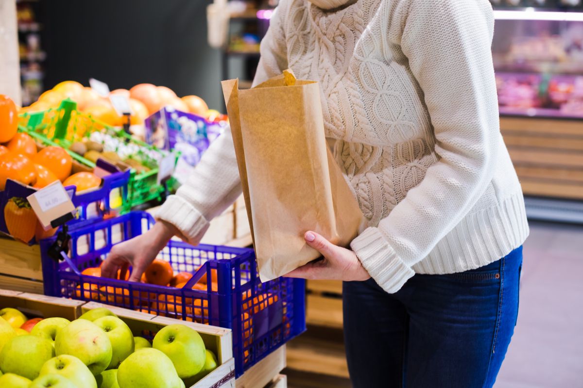 The photo shows fruits displayed and a girl holding a paper bag putting them inside indicating that buying in bulk helps avoid impulse purchases and makes it easier to save.