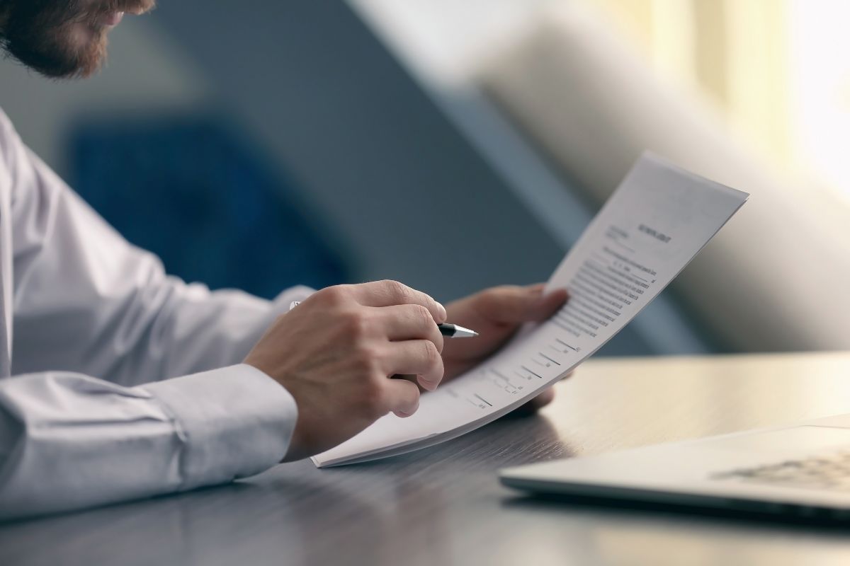 The image is about a laptop and a man holding papers and a pen referring to tracking your net worth as a motivator to keep saving and investing.