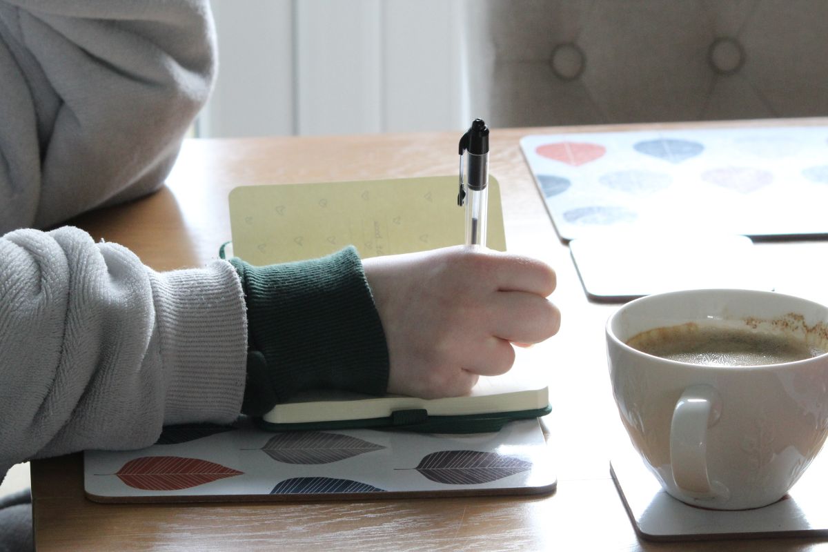 The photo shows a girl's hand and writing in the notebook while having a coffee indicating that making a money-win list helps reinforce positive money behavior and keeps you moving forward.