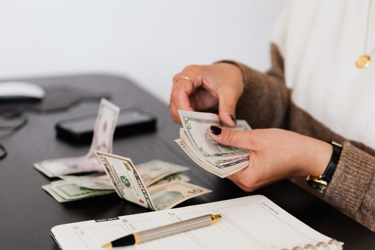 The photo tells about a notebook, a pen, dollar bills, and a girl grouping money which refers to creating a buffer for fluctuating expenses making sure the budget stays balanced even when expenses aren't the same every month.