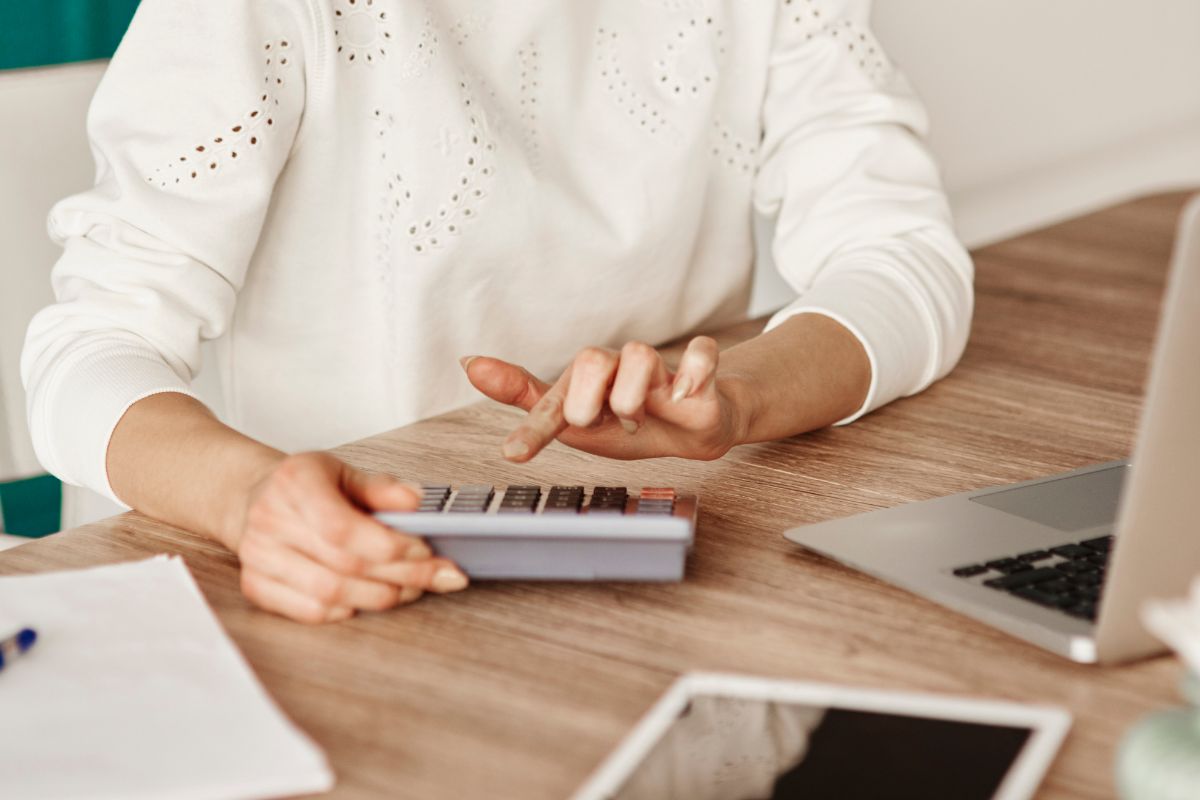 The photo shows a laptop, papers, a pen, a tablet device, and a girl's hand calculating which means reviewing and adjusting the budget monthly to keep spending in line with current goals.