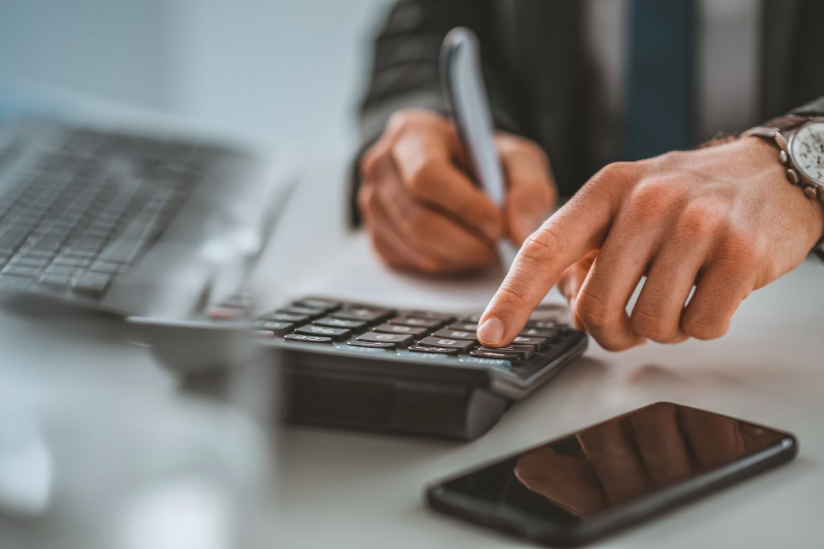 The image tells about a phone, a laptop, and a man's hand calculating while writing on paper indicating to calculate your net worth monthly helps you measure progress toward financial independence.