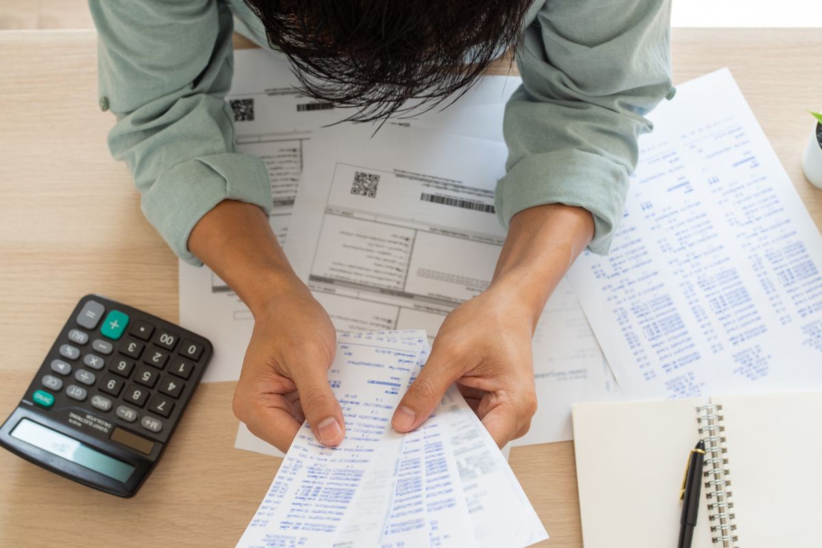 The photo shows papers, a pen, and notebook, a calculator, and a man holding receipts indicating that scheduling financial check-ins keeps you accountable and makes reaching goals easier.