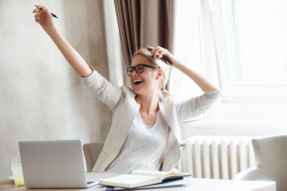 The photo tells about a notebook on the table, a laptop, and a girl cheering referring to acknowledge and celebrate small financial wins that keep you motivated to stick to good financial habits.