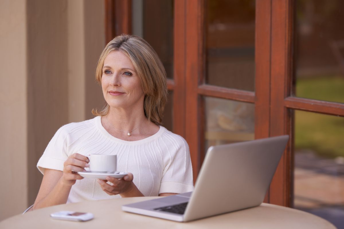 The picture of a laptop, a phone, and a girl thinking deeply while having a coffee referring to reflecting on past financial growth helps avoid past mistakes and more control you have over your finances