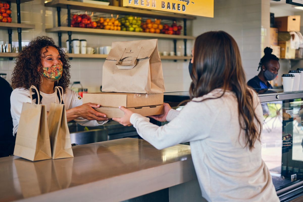 The photo tells about paper bags on the table, product display, and a cashier handing the orders in the boxes and paper bag to the customer which means to reduce takeout to save money and find budget-friendly recipes to save even more.