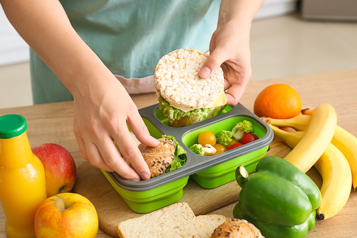 The photo tells about fruits, vegetables, a bottled drink, and a girl's hand filling the container with food which means packing lunch instead of buying at work is much cheaper and can save money.