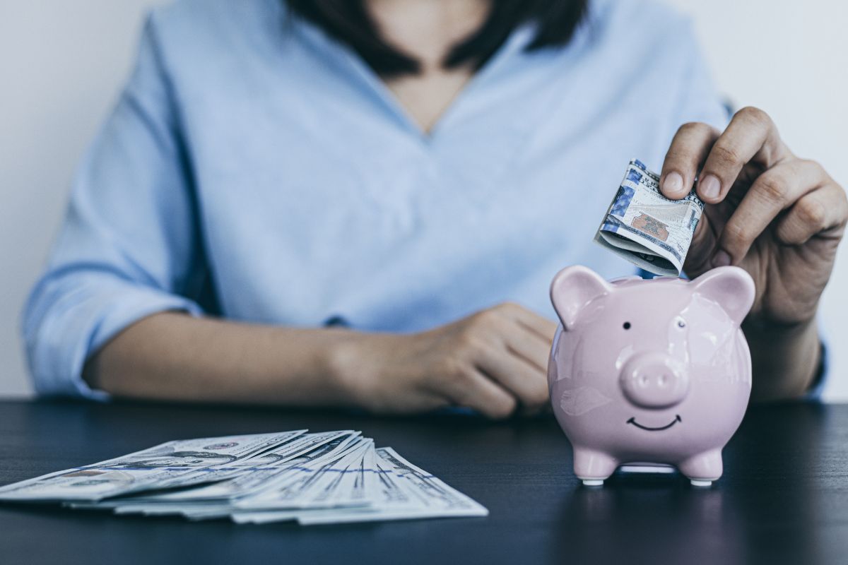 The image shows dollar bills and a girl putting a dollar bill in the piggy bank indicating to start to save 5000 in 6 months and reach your savings goals in no time.