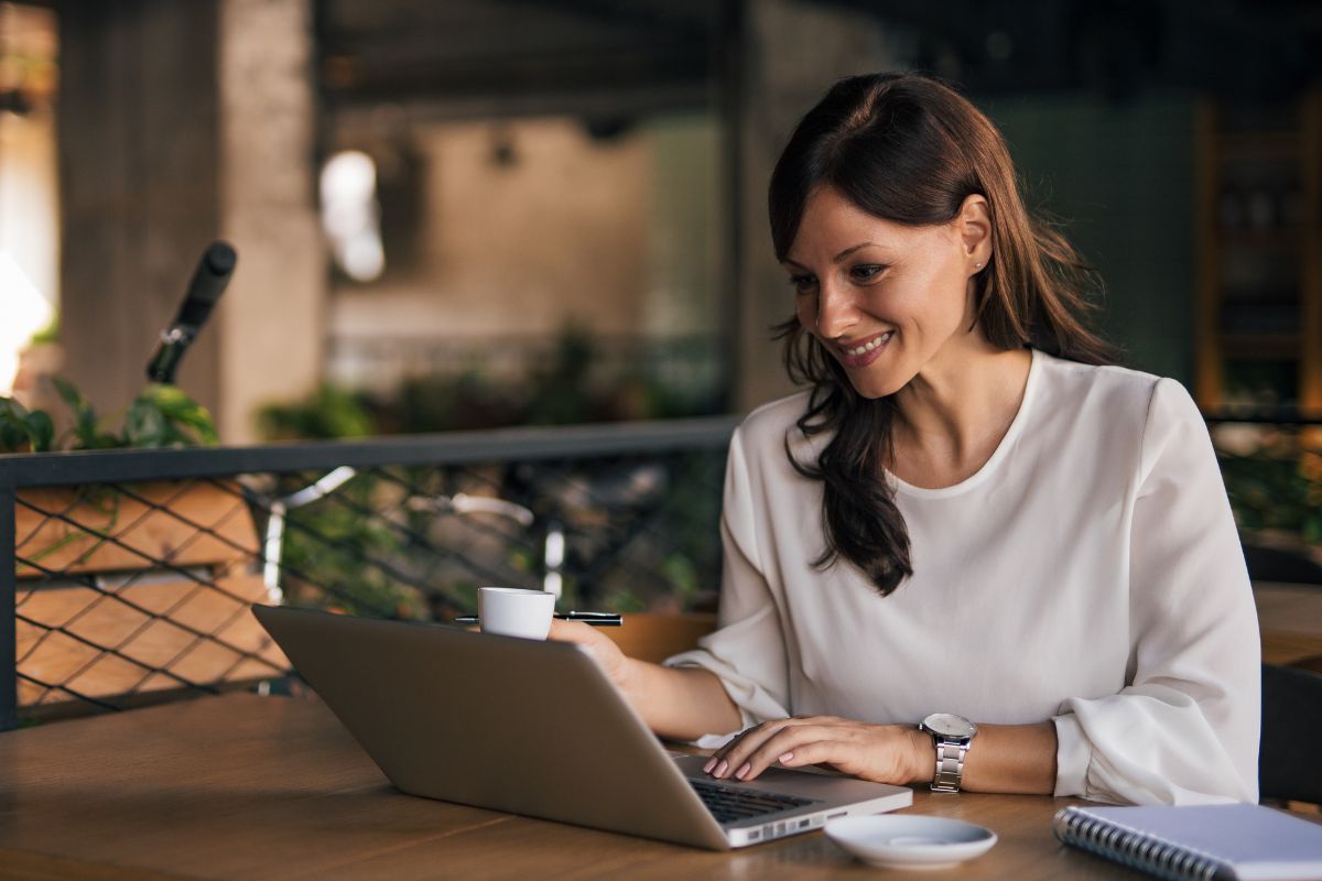 The photo tells about a notebook, a small plate, and a girl using a laptop while having a coffee referring to selling digital products that can turn creativity into a steady income.