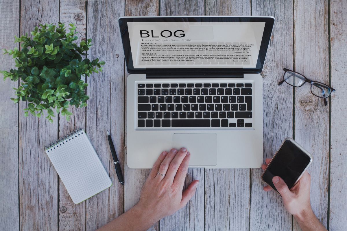 The photo shows a plant, a notebook with a pen, an eyeglass, and a man's hand using a laptop while holding a phone which refers to creating and monetizing a blog is a great option for those willing to share knowledge while creating a long-term income stream.