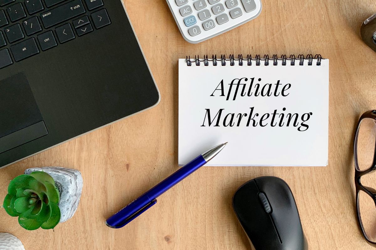 The picture tells about a laptop, a calculator, a plant, a mouse, an eyeglass, and a pen with a notebook and notes referring to affiliate marketing through social media as a simple way to monetize content while helping others find useful products.