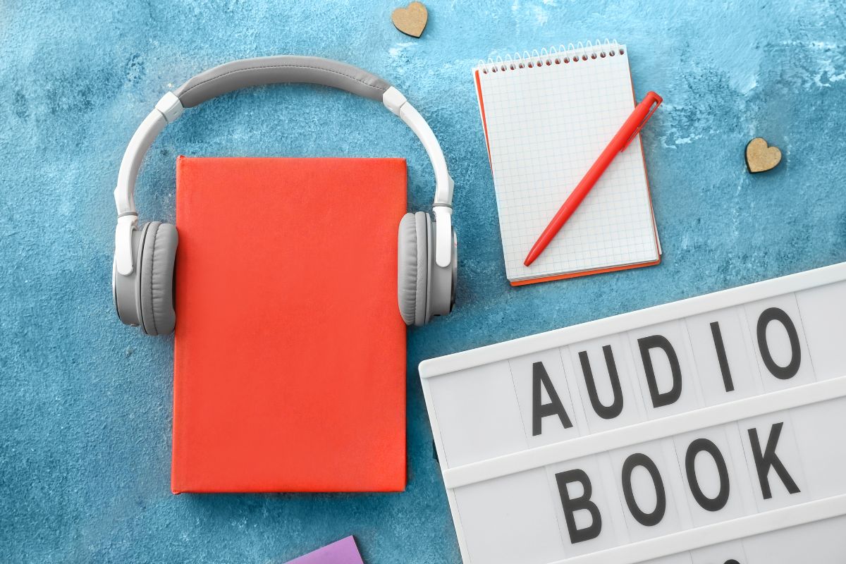 The photo shows a notebook with a pen, an earphone with a book, and a whiteboard with notes which means that creating and selling audiobooks is a good way to make passive income.
