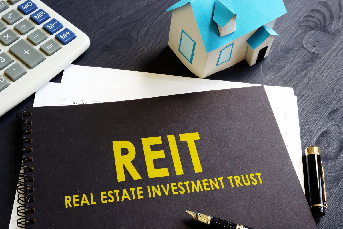 The picture shows a calculator, a mini house, a pen, papers, and a black notebook cover with notes referring to investing in REITs as an easy way to earn without managing tenants.