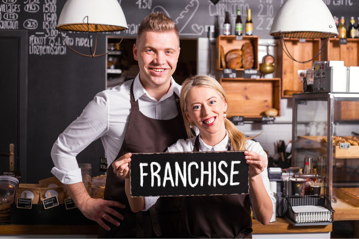 The picture tells about lamps, wooden stands, store stuff, and a girl and boy with their apron holding a signage referring to investing in franchises for semi-passive revenue can provide consistent income without the effort of building a business from the ground up.