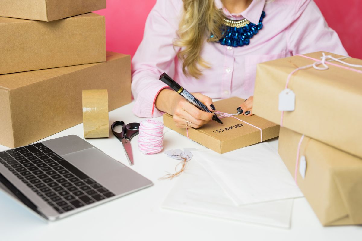 The image shows boxes, a laptop, a tape, a scissor, a yarn, and a girl's hand and writing on the box which means selling unwanted items on platforms is a quick way to make money from home.