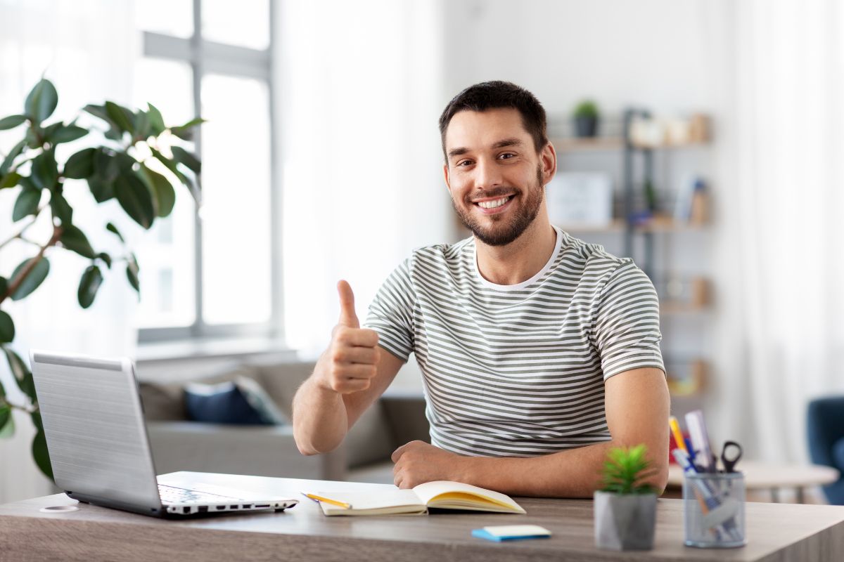 The picture shows a laptop, a notebook, a plant, pens, and a man doing an ok sign which refers to freelancing that lets you make money online using skills with flexible time.