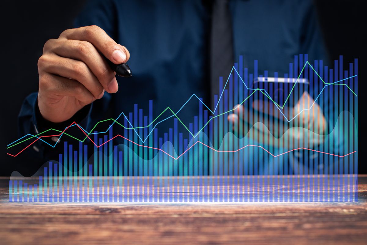 The picture tells about a man holding a phone and pen with a visual graph on the screen indicating that trading stocks is a way to grow wealth.