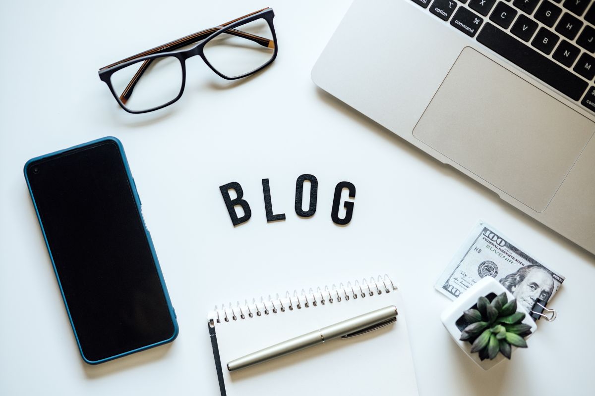 The image shows a phone, a laptop, an eyeglass, a pen, a plant, dollar bills, and a letter on the table referring to blogging for ad revenue as a way to build wealth without a traditional job.