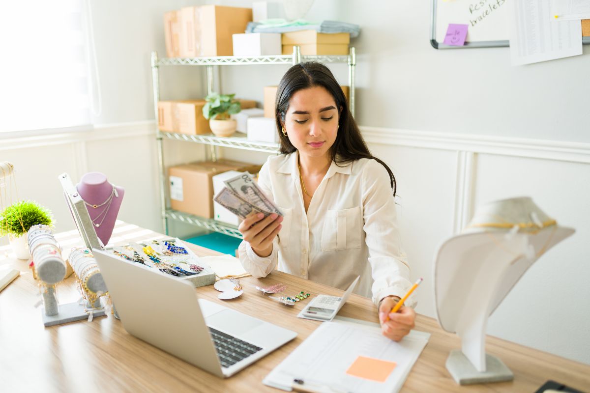 The photo shows boxes, jewelry, a laptop, and a girl writing on the paper while holding dollar bills which means running an Etsy shop for handmade is a side hustle to work from home and build financial independence. 
