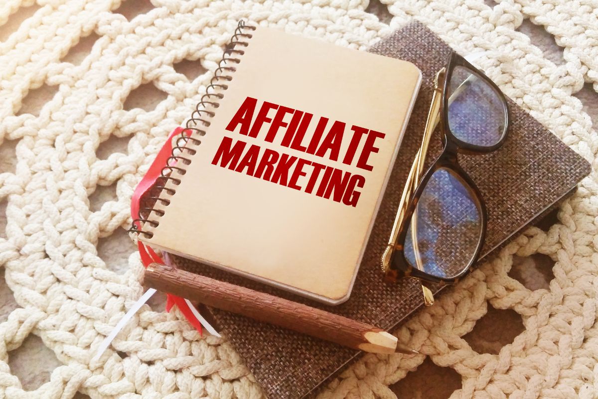 The image shows an eyeglass, a pen, and brown notebook, and a mini notebook with notes indicating that affiliate marketing through social media makes passive income as their content gets more views.