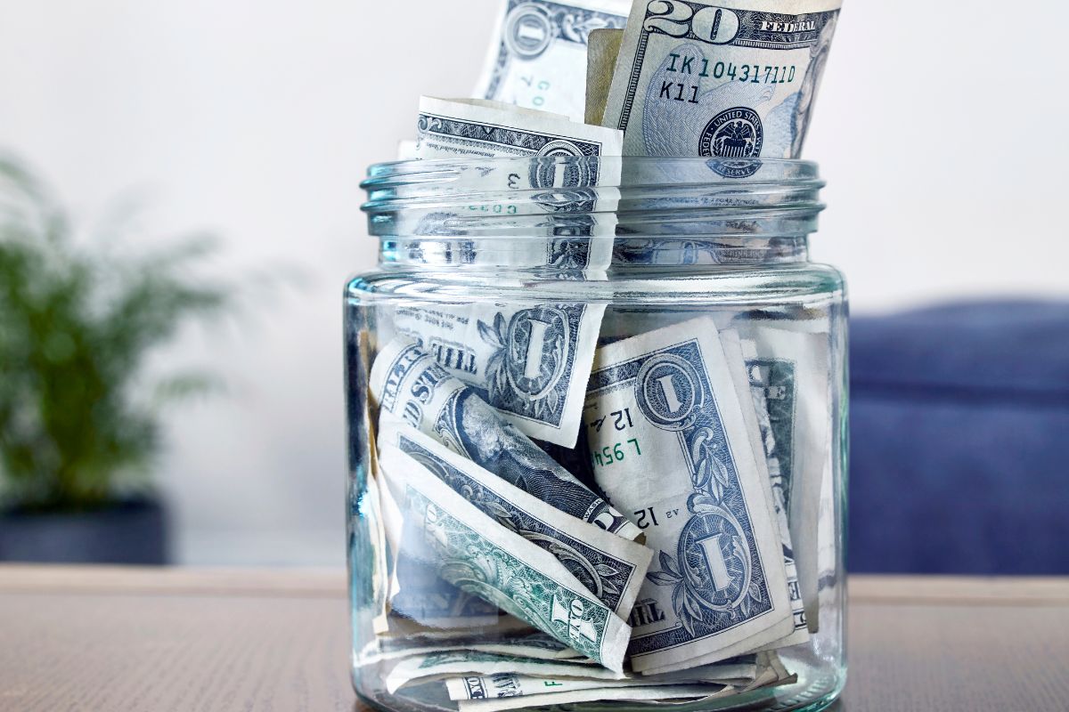 The image shows ajr filled with dollar bills indicating savings for a buffer for unexpected expenses can prevent big problems later and keep financial stress low.