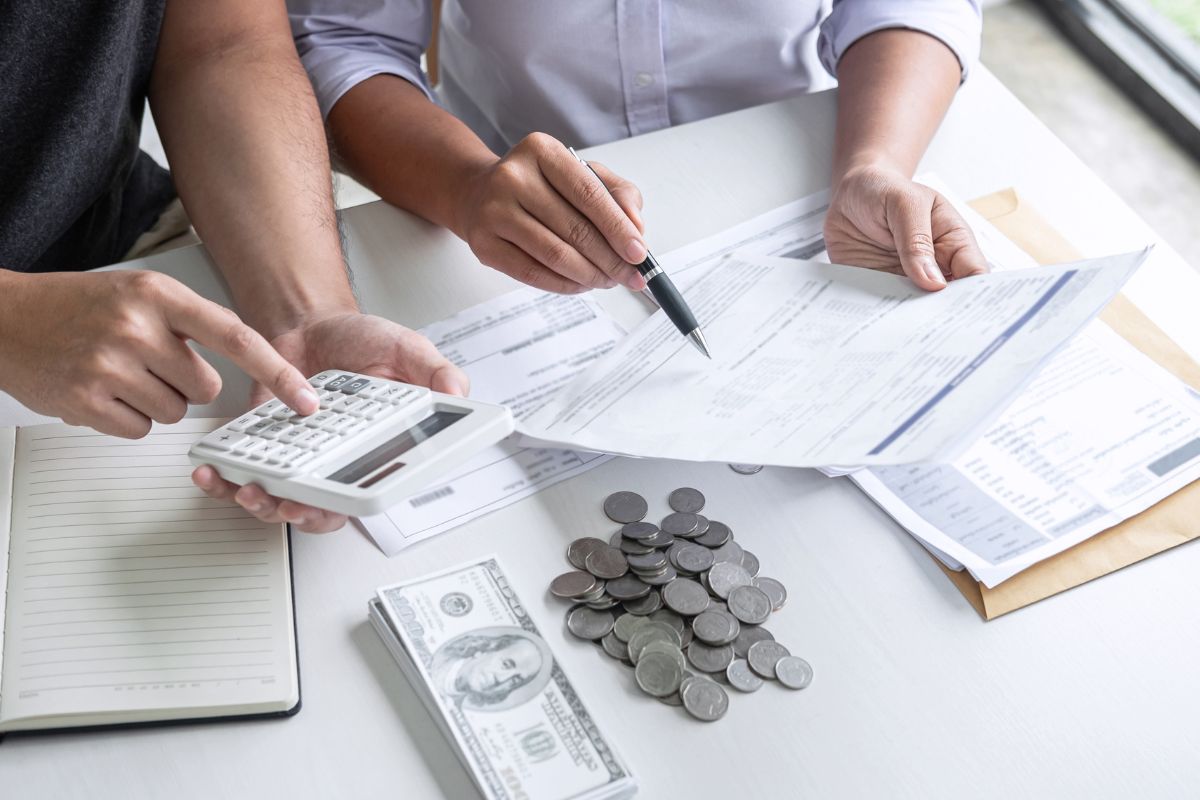 The image tells about coins, dollar bills, a notebook, papers, a man calculating, and a girl pointing the paper with a pen referring to what typically covers 3-6 months of essential living expenses means staying afloat without borrowing.