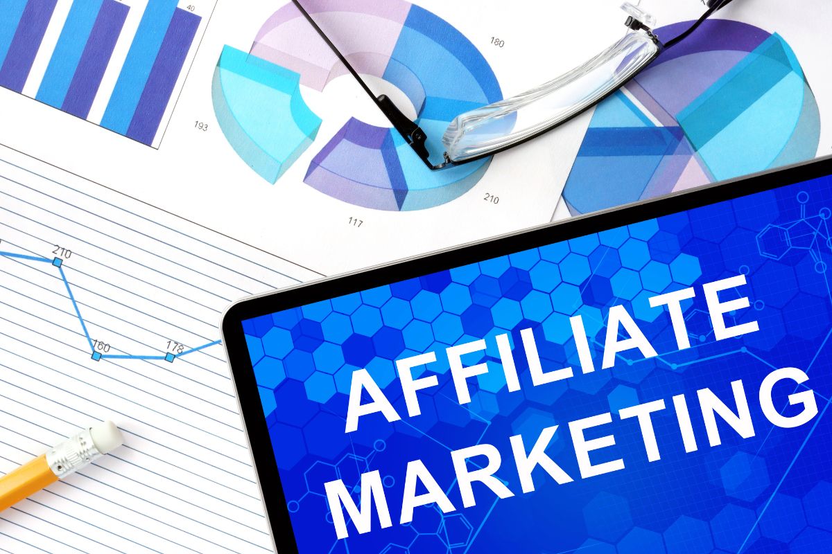 The image tells about an eyeglass, a pen, a paper with notes and graphs, and a tablet device with a screen display which refers to affiliate marketing through online platforms as a simple way to make money by recommending products online.