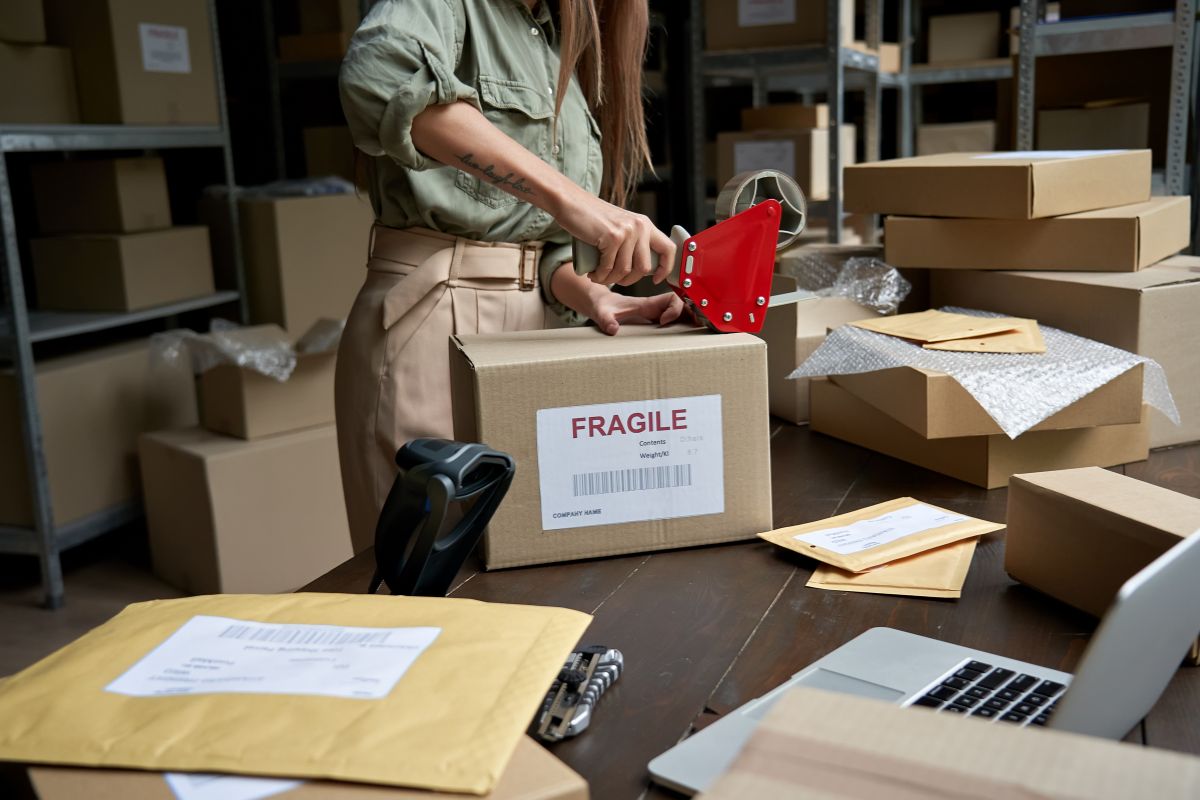 The picture tells about boxes at the warehouse, a cutter, a laptop, and a girl taping the box with a note referring to Dropshipping with Shopify is a good way to start an online business with little money.