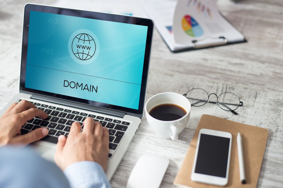 The picture tells about a clipboard, coffee, an eyeglass, a phone, a pen, a mouse, and a man's hand using a laptop with a screen display indicating that flipping domain names can be a way to make extra cash and learn to spot good deals.