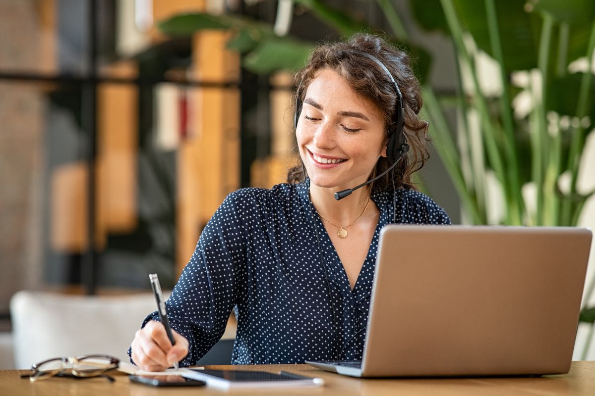 The picture shows an eyeglass on the table, a phone, a notebook, and a girl using a laptop and writing on the paper which refers to remote customer service as a steady income job that doesn't require special skills.