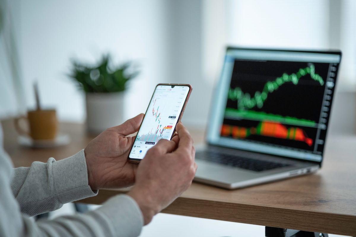 The image tells about a laptop with a screen display, a plant, coffee, and a man's hand using a phone with a screen display indicating that trading stocks or options can be a way to make money online if done carefully.