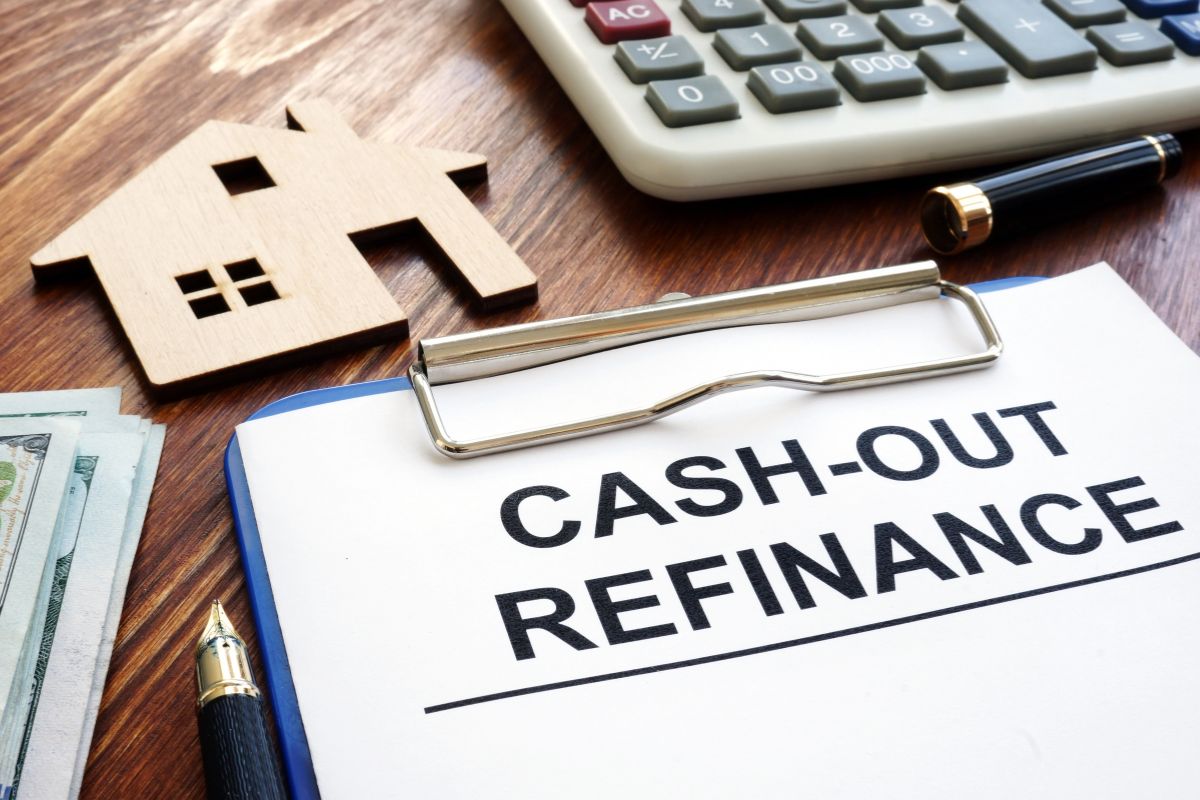 The photo tells about a calculator, a wooden mini house figure, dollar bills, a pen, and a clipboard with notes indicating that taking out cash with a refinance can be helpful for paying off interest debt of funding home improvements.
