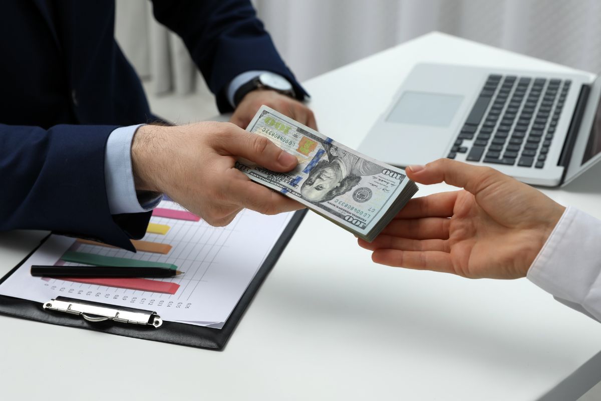The photo tells about a laptop, a clipboard with a graph, a pen, and a man in a suit handing the buck of dollar bills to the man's hand indicating that comparing offers from multiple lenders help you find the best deal and avoid overpaying.