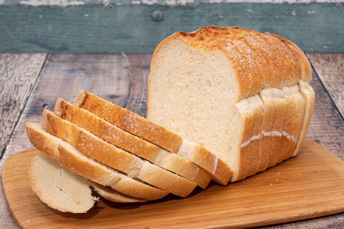 The image shows a slice of bread on a wooden board referring to white bread and refined grains are cheap but don't provide much nutrition, whole bread makes meals more filling and stretches a grocery budget further.