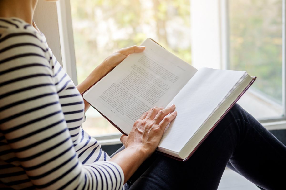 The picture shows a girl reading a book beside the window referring to finding free entertainment options that can help you save money while still enjoying.