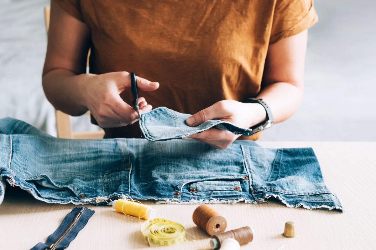 The image shows sewing yarns, tape measure, a denim clothe, and a girl cutting the fabric which means repairing and repurposing items can save money and keep things out of trash.