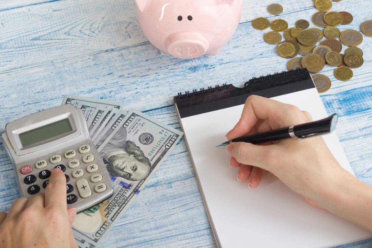 The image shows coins, a piggy bank, dollar bills, and a girl's hand calculating and writing on paper referring to creating a realistic budget that supports your financial independence without feeling impossible to stick to.