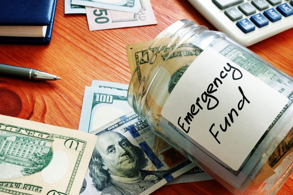 The image tells about a calculator, a notebook, dollar bills, and a jar filled with dollar bills with a note indicating that building an emergency fund can keep you from going into debt when the unexpected happens.