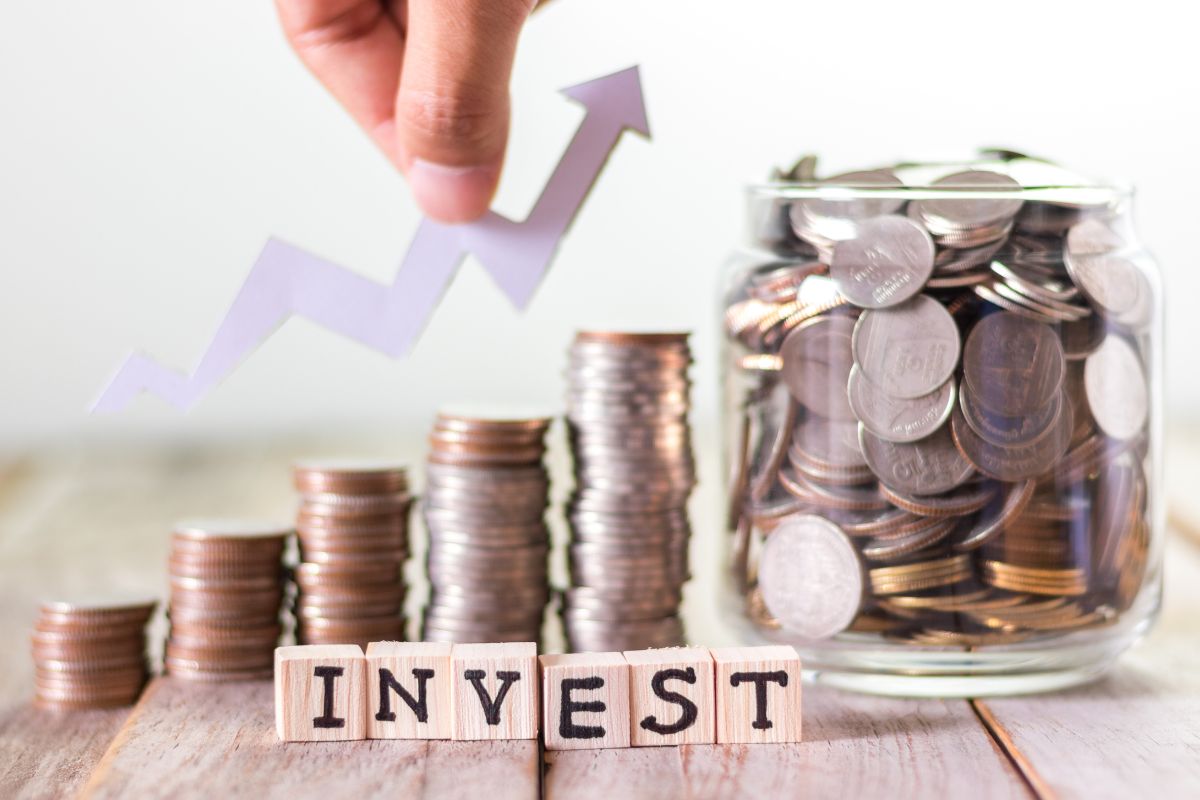 The picture shows a jar filled with coins, stock coins, wooden cubes with letters, and a man's hand holding an arrow which means investing consistently in diverse assets can bring financial stability and give you more freedom in the future.